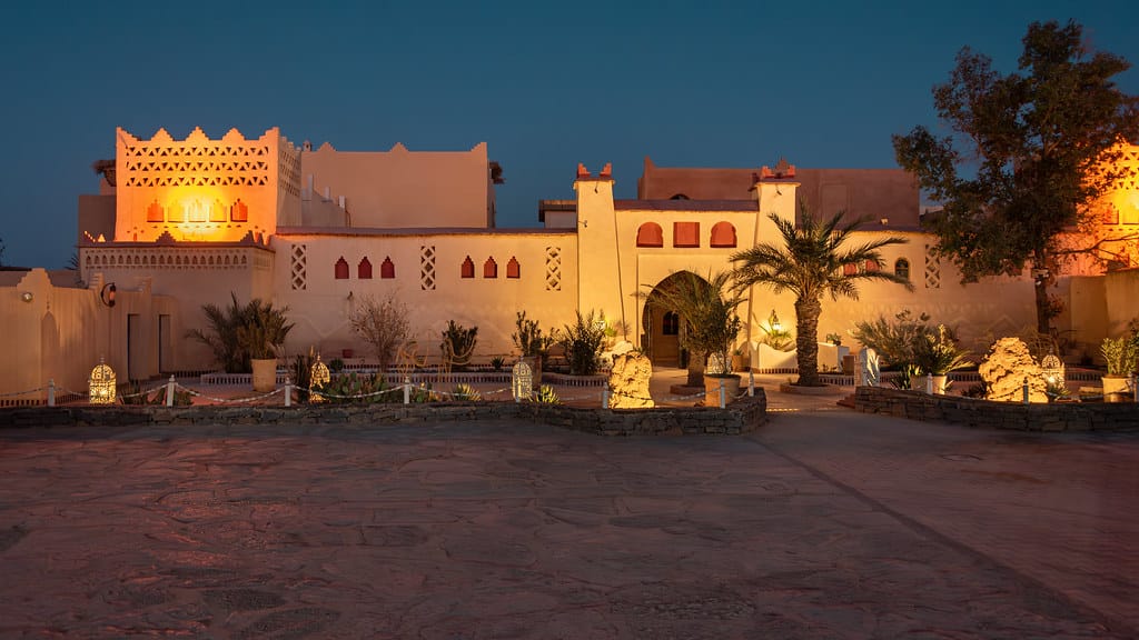 Illuminated traditional Moroccan kasbah with golden lighting, palm trees, and decorative lanterns in the garden at night